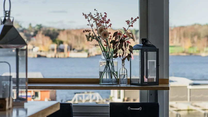A glass vase with flowers and a black lantern sit on a windowsill, overlooking a calm waterfront with blurred trees in the background.