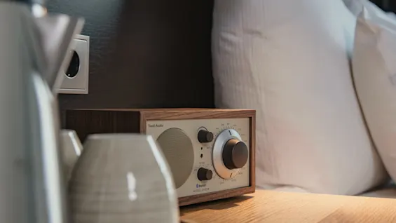A bedside table holds a kettle, cups, and a vintage radio under a wall-mounted reading lamp; pillows rest against a dark wooden headboard, creating a cozy bedroom setting.
