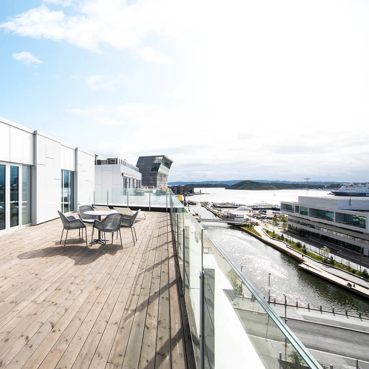 A rooftop terrace with wooden flooring features a table and chairs. It overlooks a modern cityscape and waterfront under a bright sky.