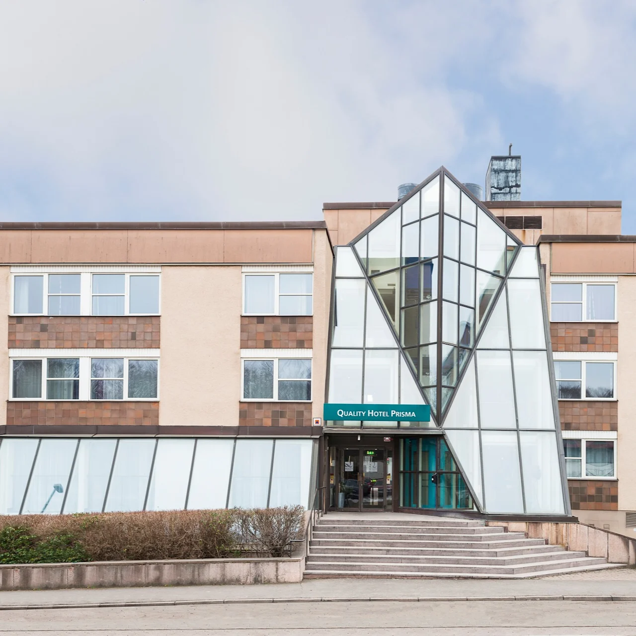 En modern hotellentré med glasfasad och geometrisk design, märkt "Quality Hotel Prisma," omgiven av buskar och ett stenbelagt område under en delvis molnig himmel.