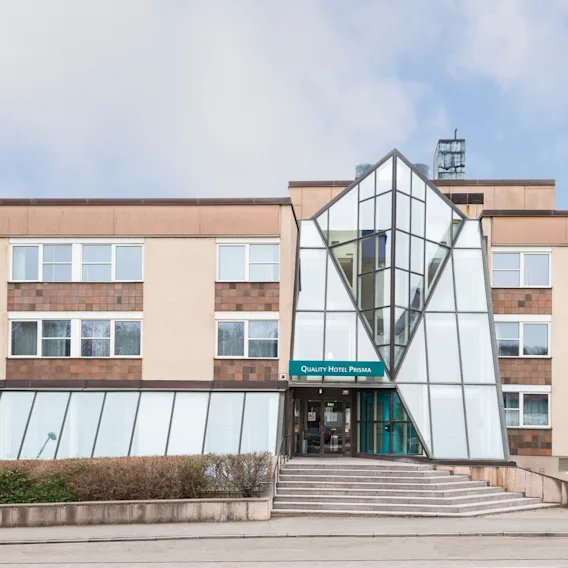 En modern hotellentré med glasfasad och geometrisk design, märkt "Quality Hotel Prisma," omgiven av buskar och ett stenbelagt område under en delvis molnig himmel.