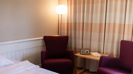To vinrøde stole er placeret omkring et lille træbord med en bog og en retro radio. En tændt gulvlampe oplyser det hyggelige rum med stribede gardiner og et sengehjørne.