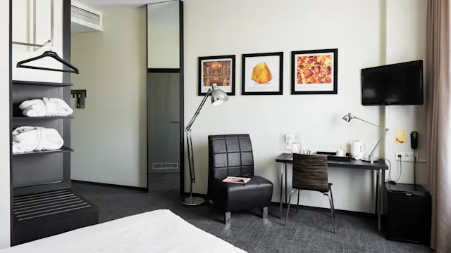 A hotel room features a modern chair beside a lamp, near a wall with three framed artworks. A desk holds various items, opposite a small television on the wall.