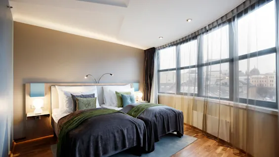 A neatly made double bed rests against an accent wall, illuminated by bedside lamps, within a modern room featuring wooden flooring and large windows overlooking a cityscape.