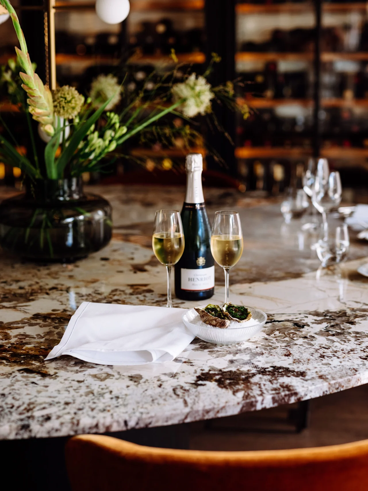 A bottle of sparkling wine and two filled glasses sit on a marble table beside a dish with oysters. A floral centerpiece and wine racks decorate the background.