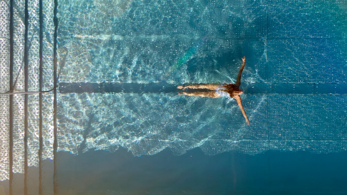 Flyfoto av en person som svømmer i et klart blått basseng med trapper, omgitt av fliser.