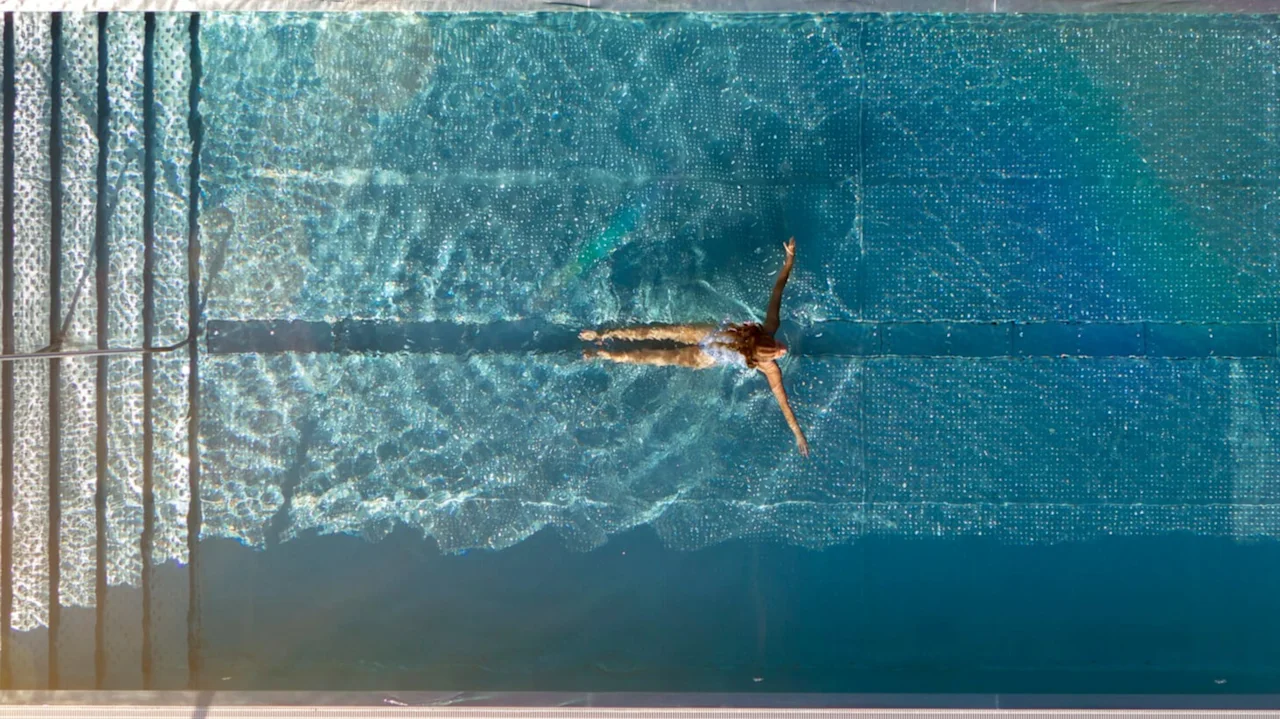 Flyfoto av en person som svømmer i et klart blått basseng med trapper, omgitt av fliser.