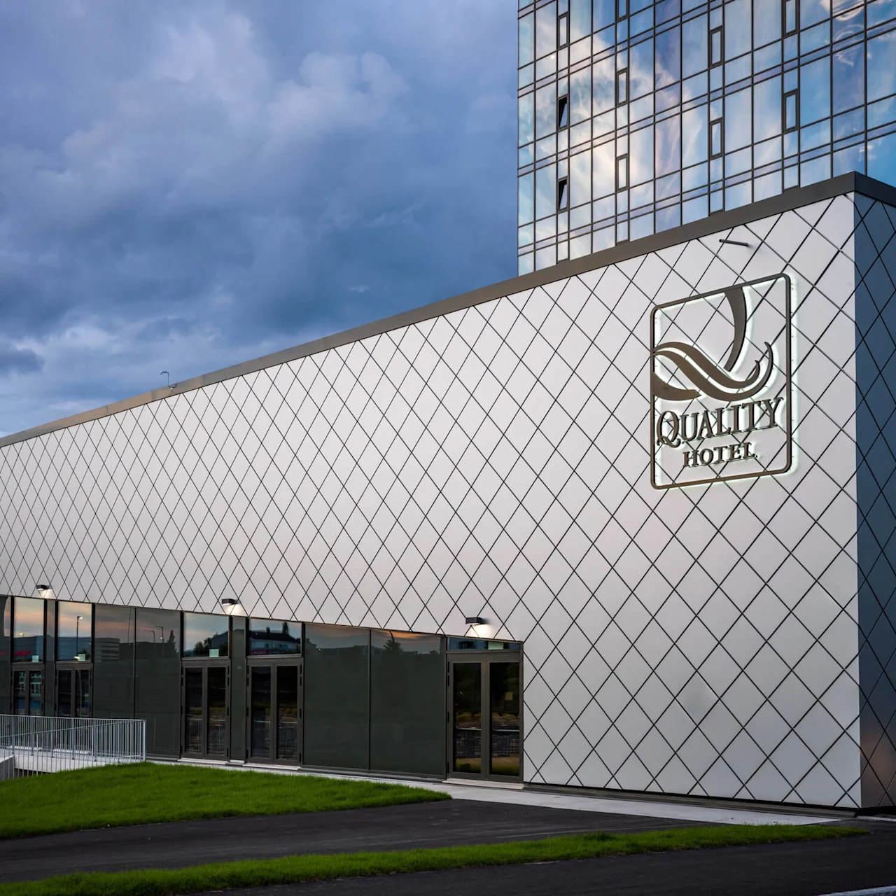 En moderne bygning med en diamantmønstret fasade har teksten "QUALITY HOTEL." Store glassdører på nederste nivå, omgitt av gressplen, under en skyet himmel.