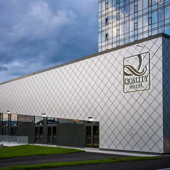 En moderne bygning med en diamantmønstret fasade har teksten "QUALITY HOTEL." Store glassdører på nederste nivå, omgitt av gressplen, under en skyet himmel.