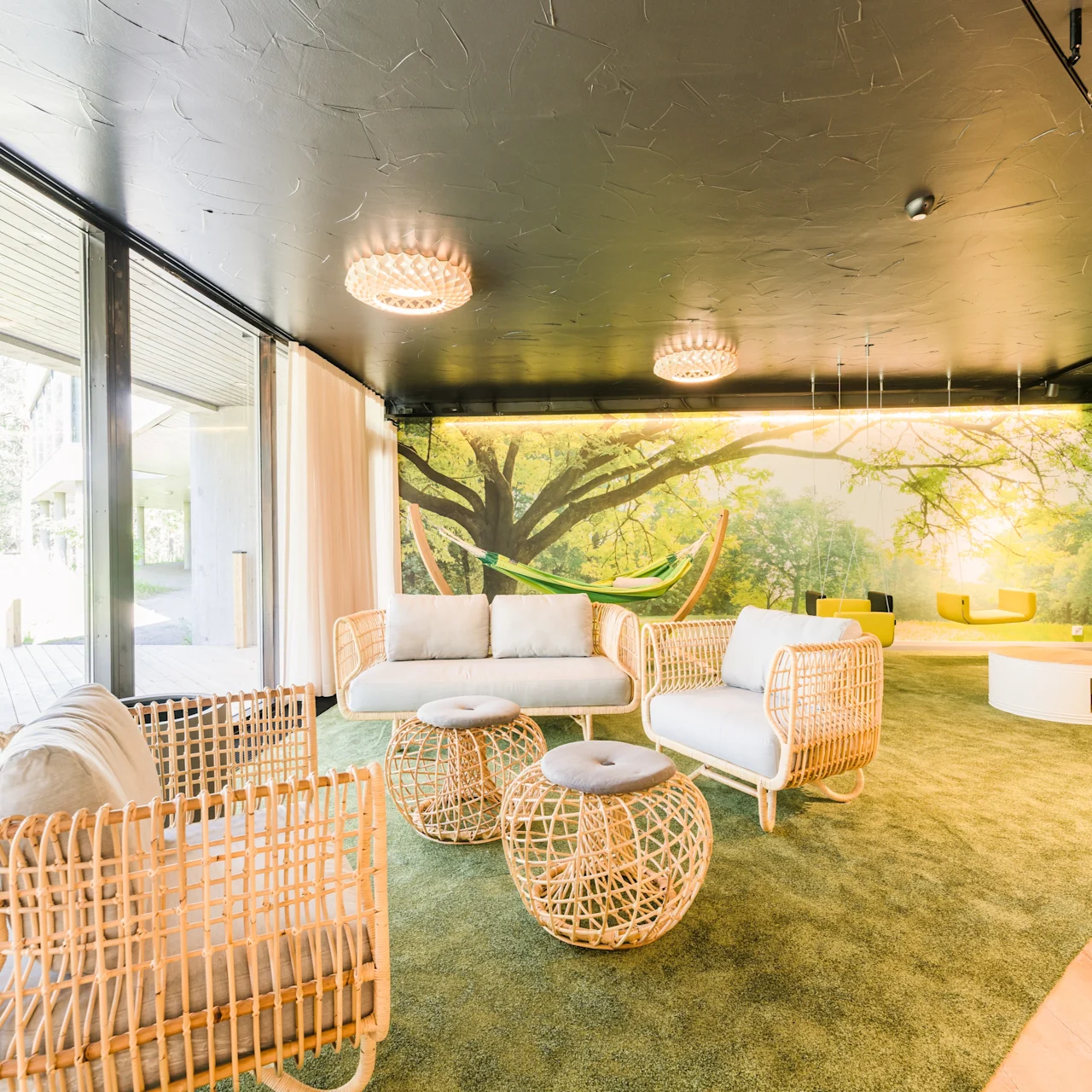 Woven furniture arranged for seating in a bright room with large windows and nature-themed wall mural, illuminated by ceiling lights, set on green carpet with outdoor view.