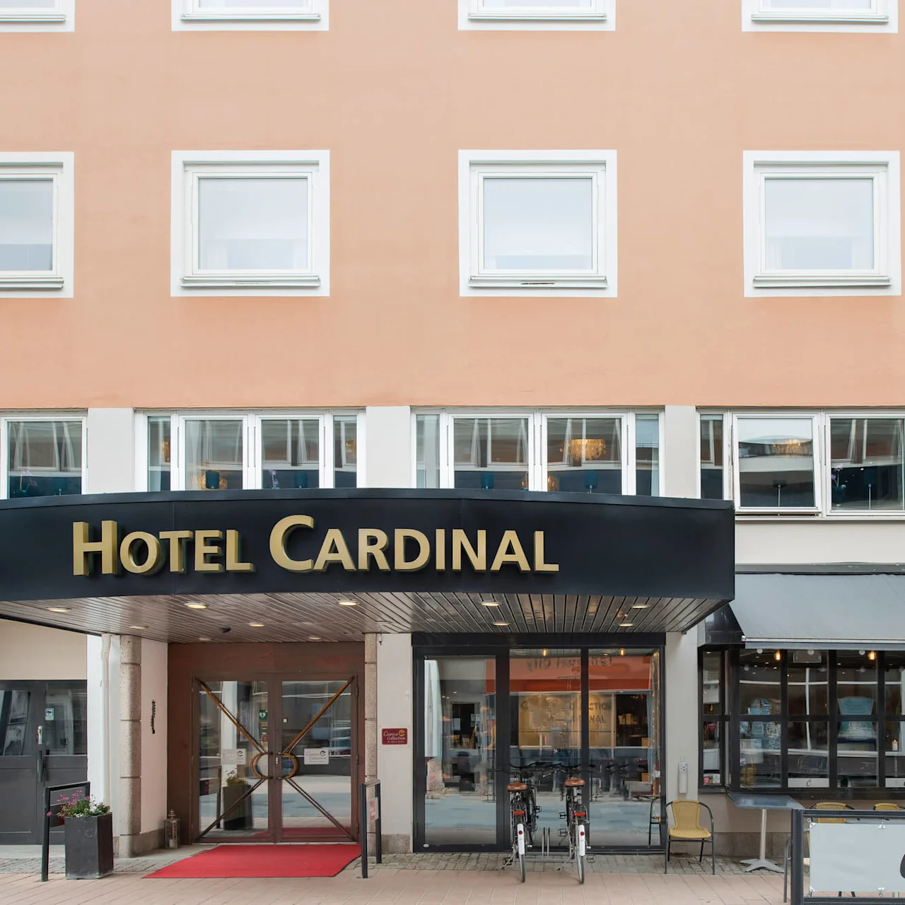 Vaaleanpunainen rakennus, jossa kyltti 'Hotel Cardinal', lasiovi alla ja polkupyörä parkkeerattuna edessä.