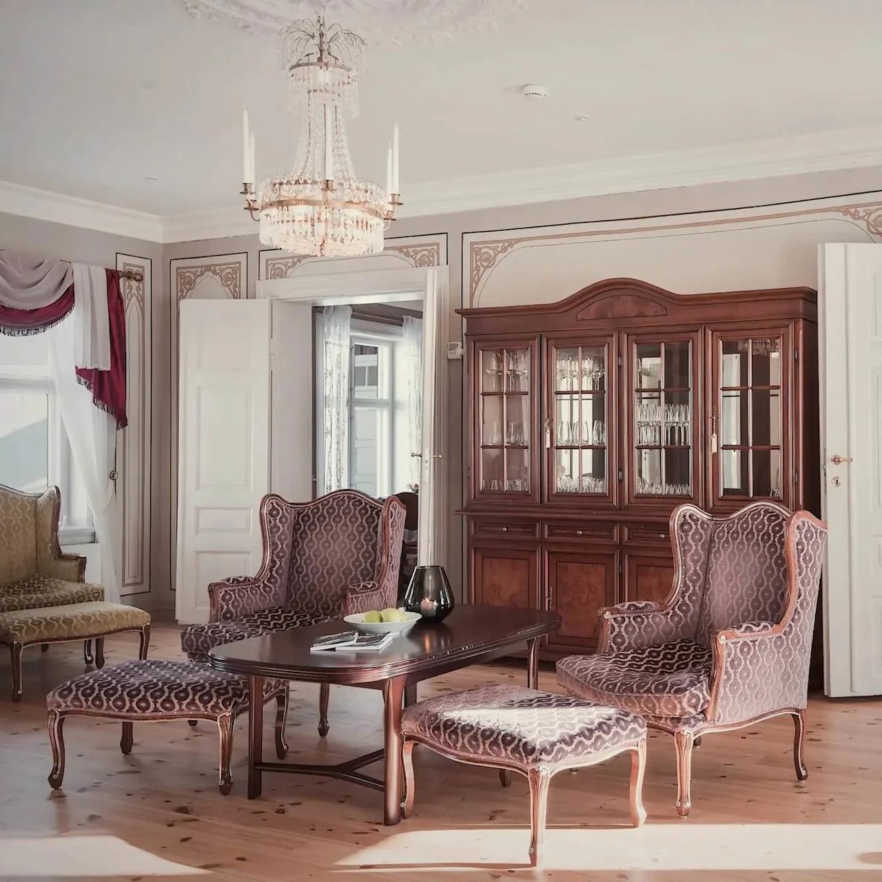 Antikt træskab står elegant, viser glas, ved siden af mønstrede stole og et centralt bord i en raffineret stue. Åbne døre fører til en spisestue dækket til et måltid.