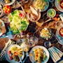Skåle og tallerkener fyldt med forskellige salater, supper og brød er arrangeret på et mørkt marmorbord, omgivet af bestik, servietter og glas, skaber en farverig middagsopsætning.