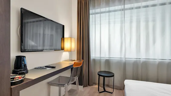 Wall-mounted TV facing a desk with a lampshade, chair, and kettle. Sheer curtains allow light through; a small round table and bed visible. Text: "CAUTION HOT WATER".