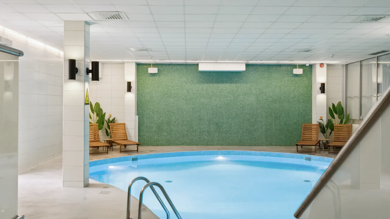 A curved indoor pool with calm water is surrounded by wooden recliners, modern lights, and potted plants. The setting is a clean, bright room with a tiled ceiling and walls.