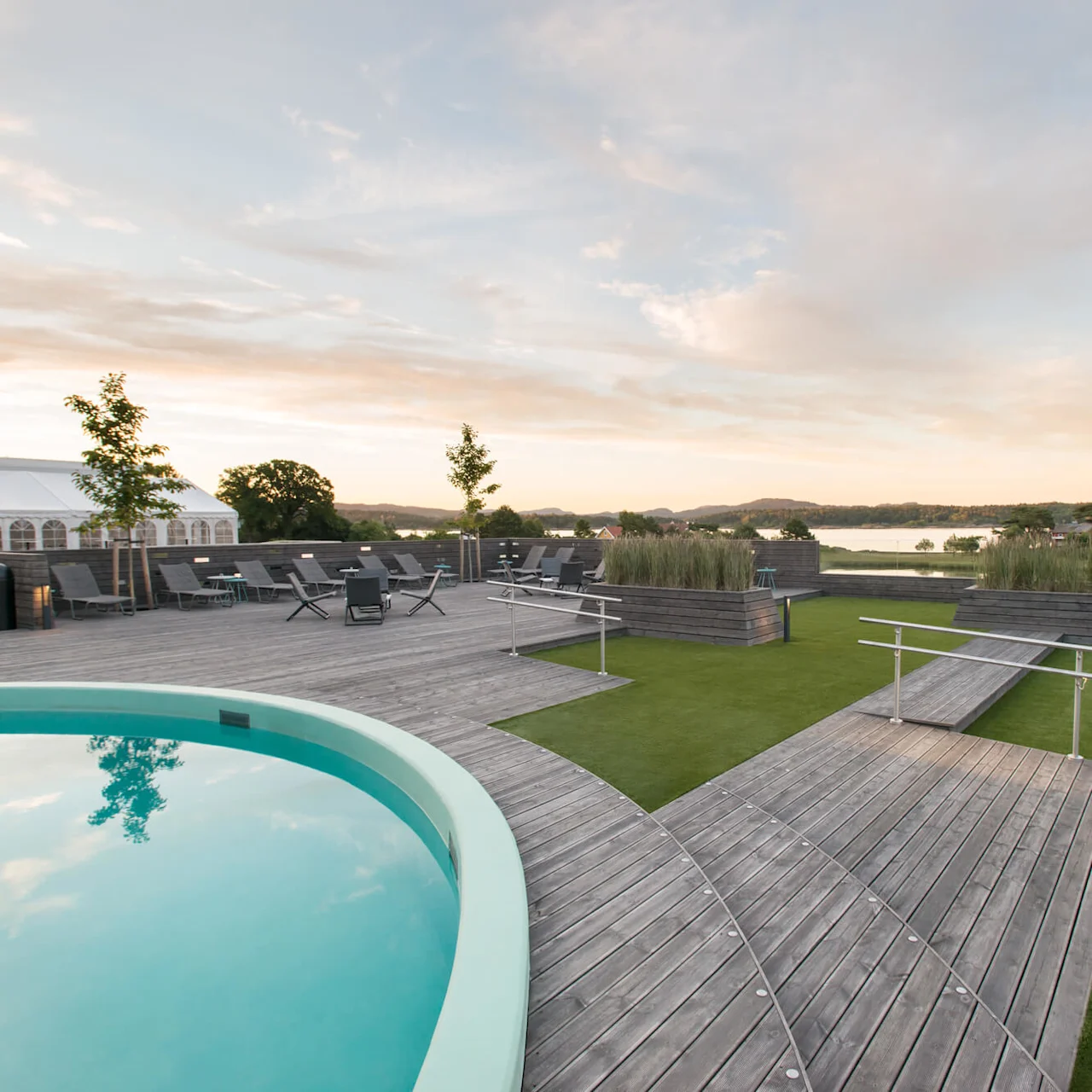 Circular pool reflecting sky, surrounded by wooden deck; chairs and trees arranged around, with a large tent nearby. Scenic landscape with hills and lake in the background at sunset.
