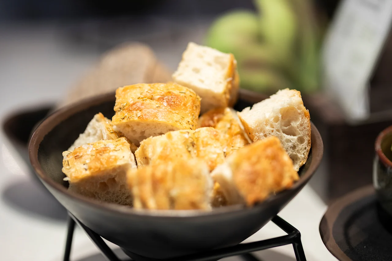 Tumma kulho täynnä kullanruskeita leipäkuutioita, ripoteltuna yrteillä ja suolalla, valmiina nautittavaksi.