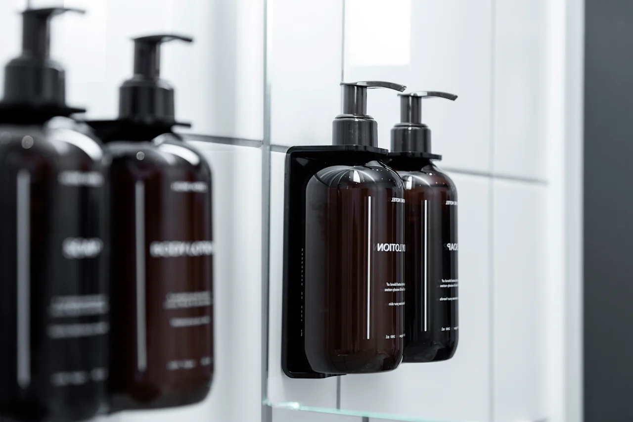 Lähikuva tummista pumppupulloista, joissa on BODY LOTION ja SHAMPOO, asennettuna valkoiselle kaakeloidulle kylpyhuoneen seinälle, heijastuen peilistä.