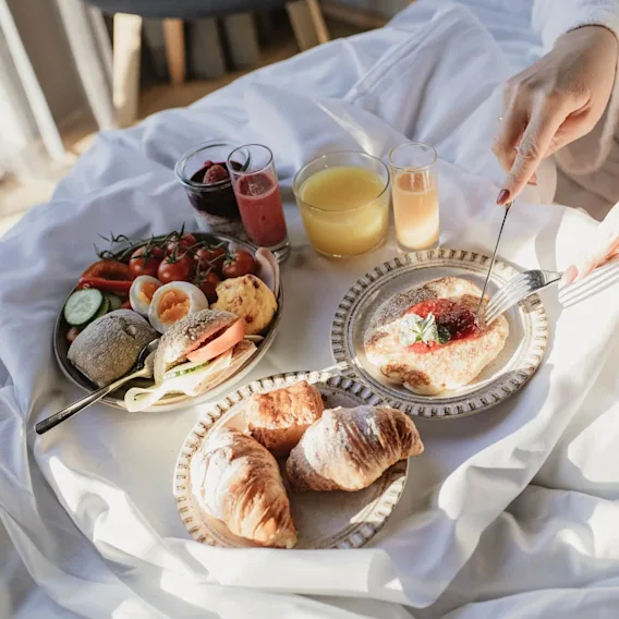 En person skjærer i en pannekake med syltetøy og mynte på, omgitt av en frokost med croissanter, juice, frukt og brød, på en seng med sollys som strømmer inn.