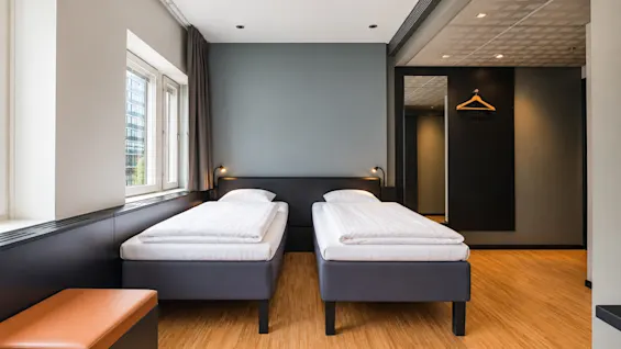 Bright hotel room featuring two single beds, wooden flooring, and a grey accent wall.