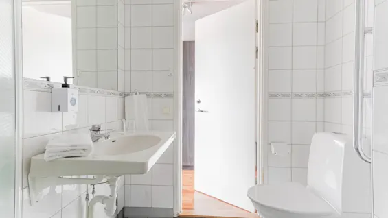 A bright, clean bathroom with white tiles, a sink, and a toilet. An open door leads to another room.