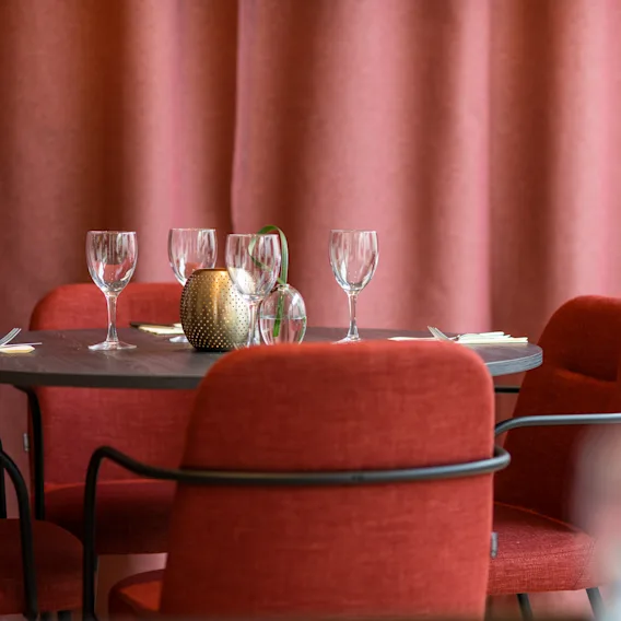 Ett restaurangområde med ett bord dukat med vinglas och röda stolar, mot en bakgrund av röda gardiner.