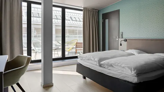 A bed with white linens rests against a teal wall, near large windows overlooking a patio with colorful chairs.