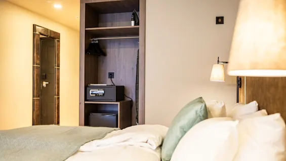 En prydligt bäddad säng med kuddar står i ett modernt hotellrum. En vägglampa lyser upp en öppen garderob med ett kassaskåp i. En dörröppning leder till ett annat område.