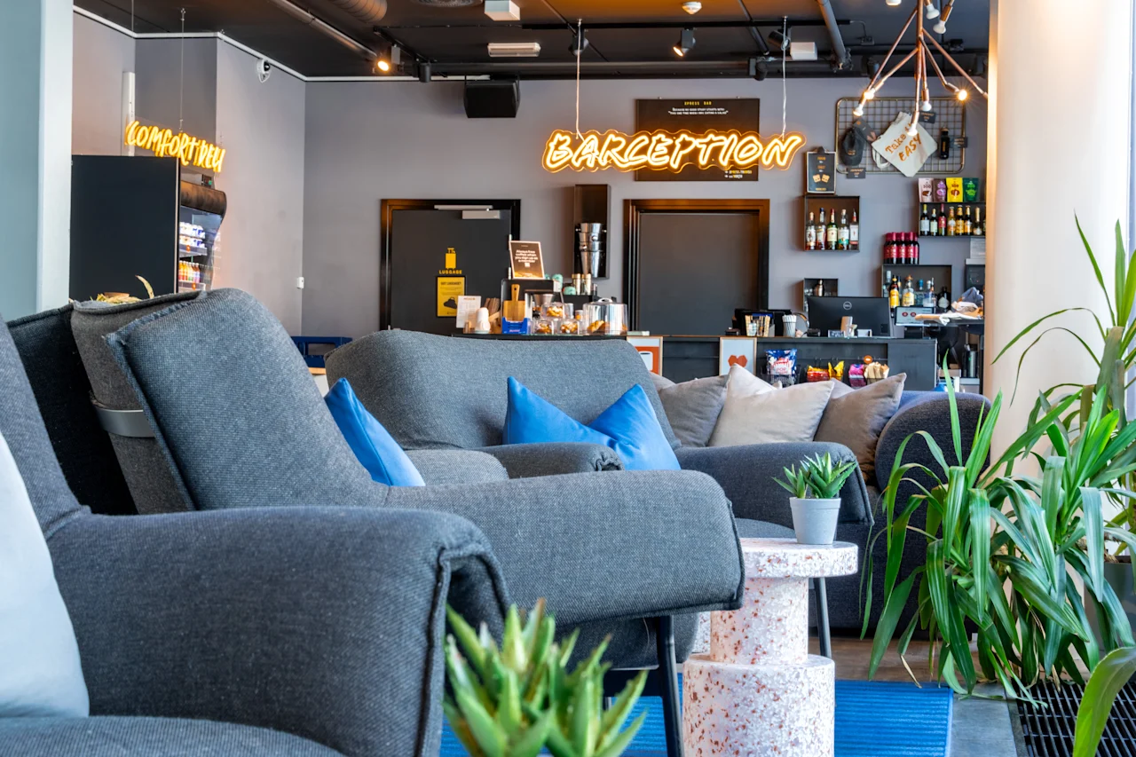 Komfortable grå stole med blå puder er arrangeret i en moderne café. Neonskilt siger "BARCEPTION" og "KOMPROVAL." Omgivelserne har planter og en velassorteret bar i baggrunden.
