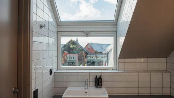 A bright, tiled bathroom with a sink, a window overlooking buildings, and a skylight revealing the sky above.