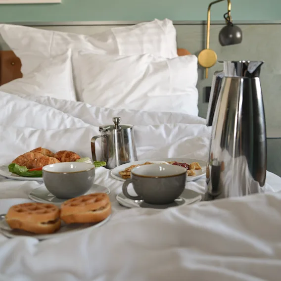 Breakfast items on a bed, including two cups, a metallic carafe, pastries, and a small teapot. The setting is a bedroom with white linens and a teal wall.