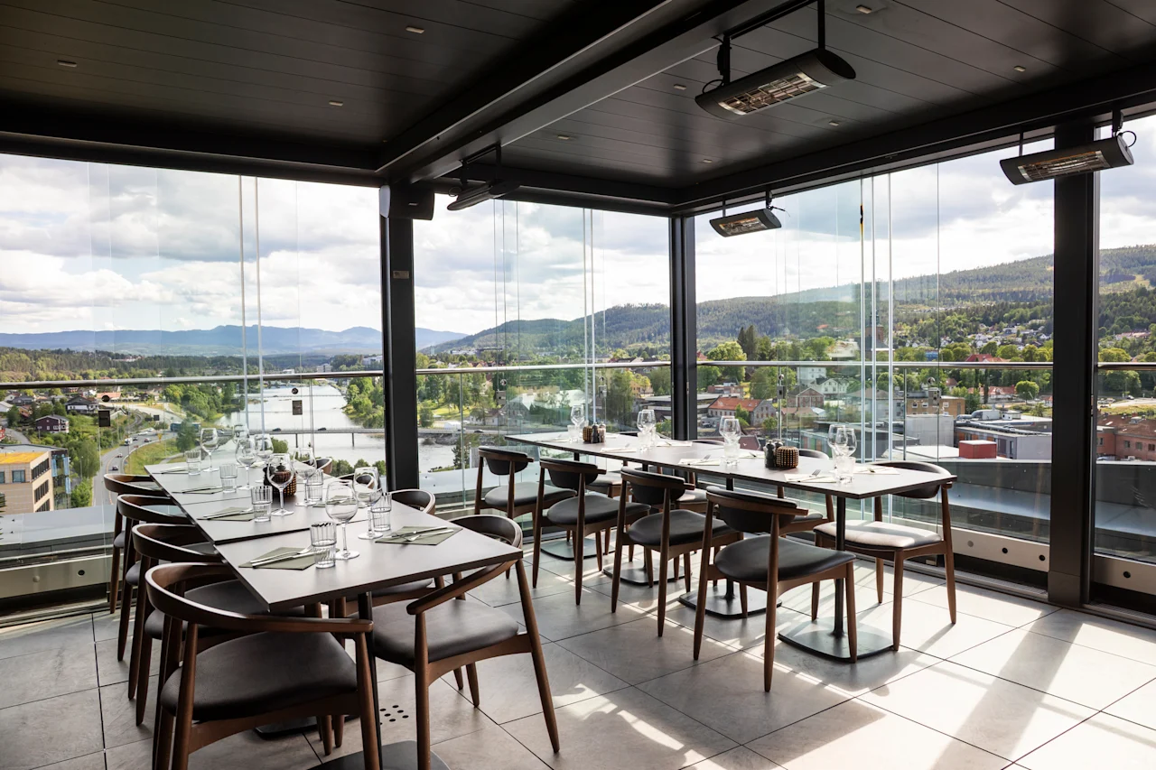 En moderne restaurant med gulv-til-tak-vinduer som tilbyr en naturskjønn utsikt over en by, elv og fjell. Bordene er dekket for bespisning.