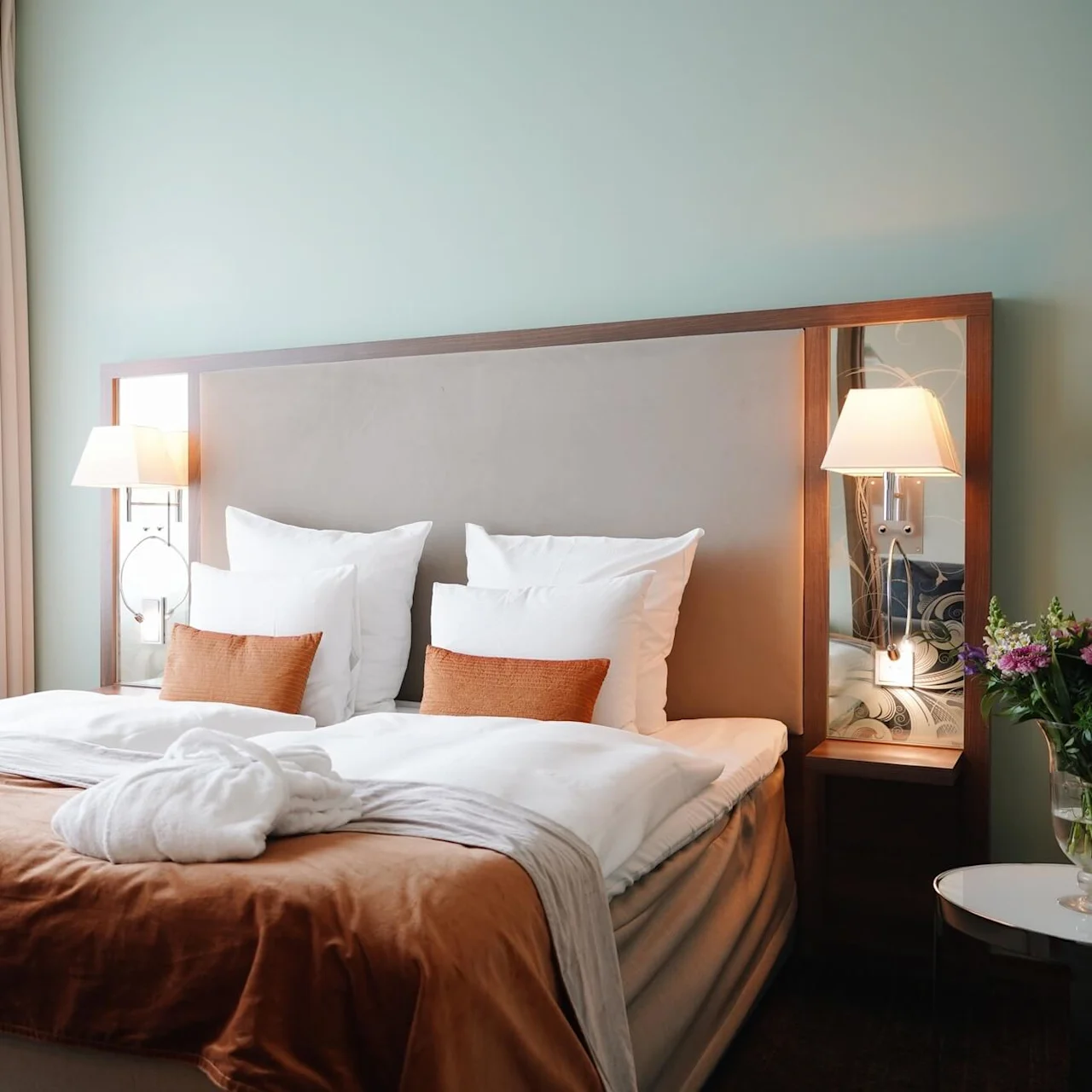 A neatly made bed with pillows and brown blanket, flanked by bedside lamps, in a softly lit, elegant bedroom featuring a vase of flowers on a side table.