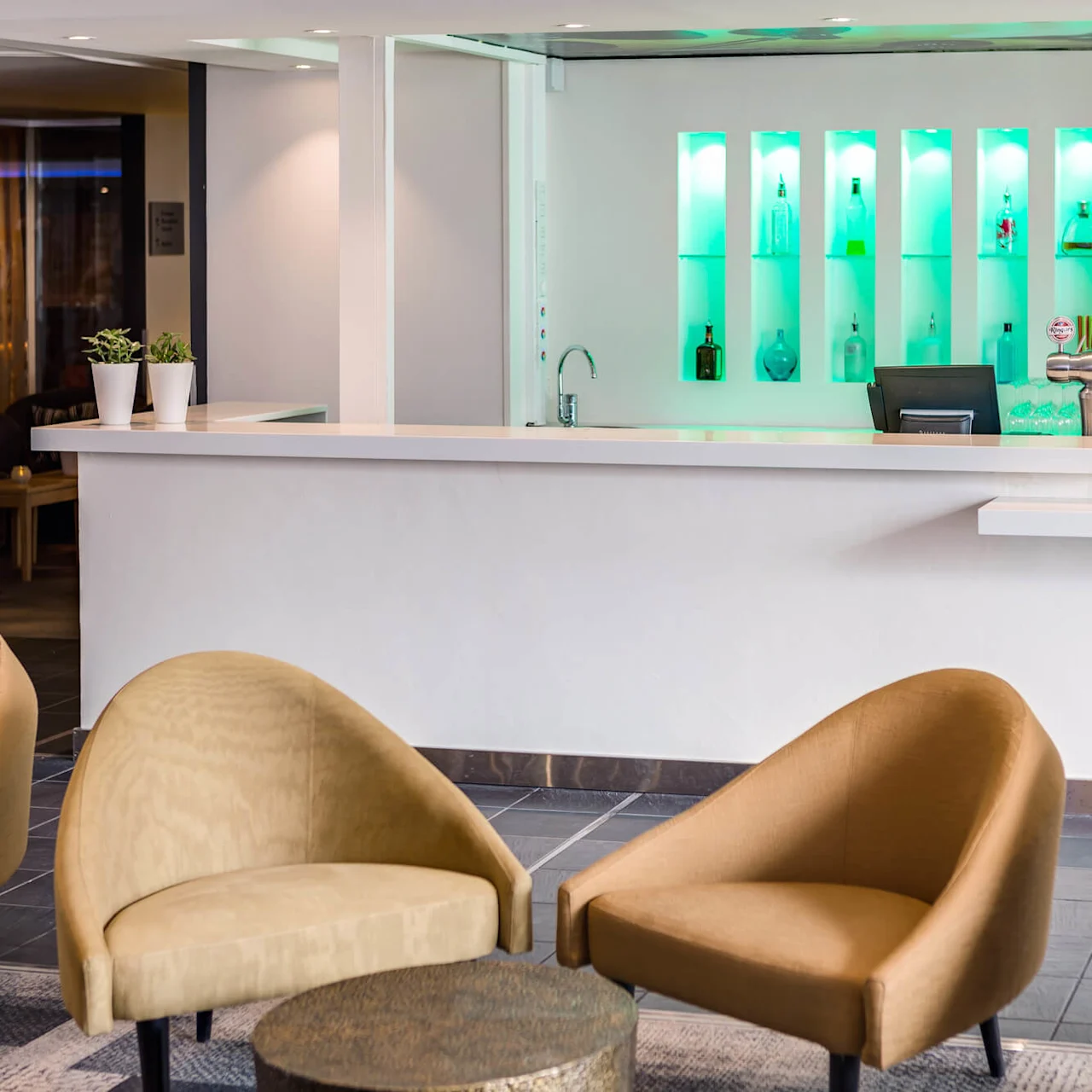 Bar counter with a sleek design features illuminated shelves holding bottles. Tan chairs are placed in the foreground, creating a modern and inviting lounge atmosphere.