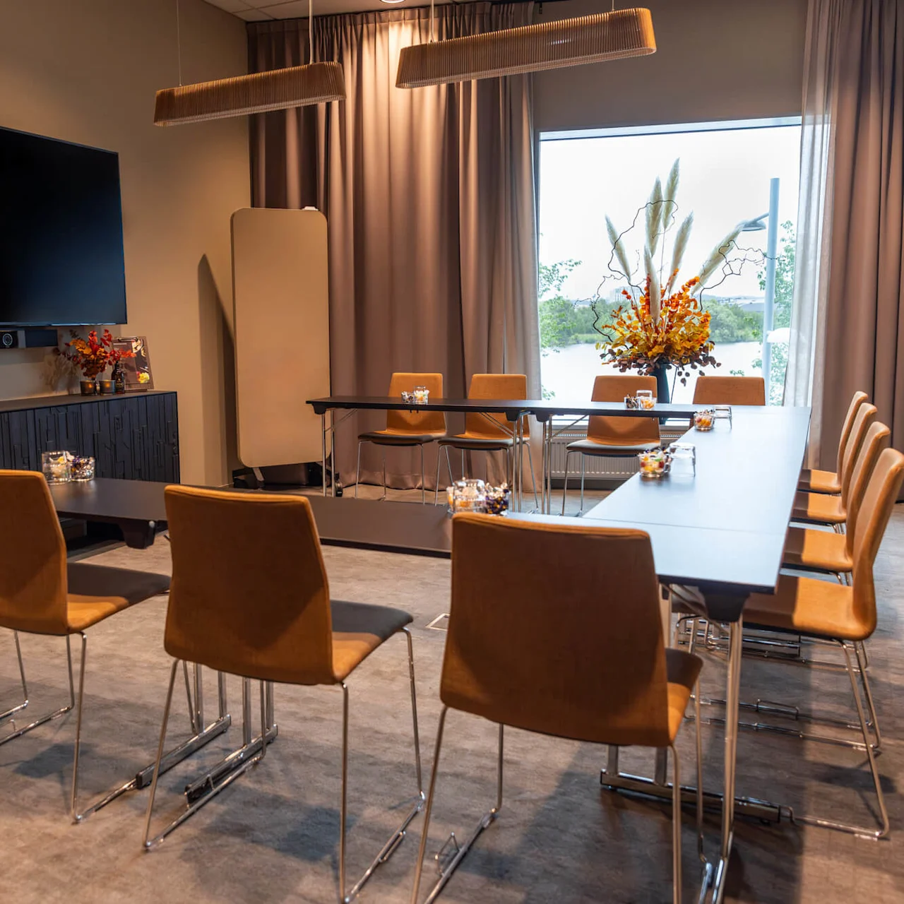 Bruna stolar runt bord, riktade mot en stor väggmonterad TV i ett mötesrum med beige väggar och ett stort fönster, dekorerat med blomsterarrangemang och överhängande belysning.