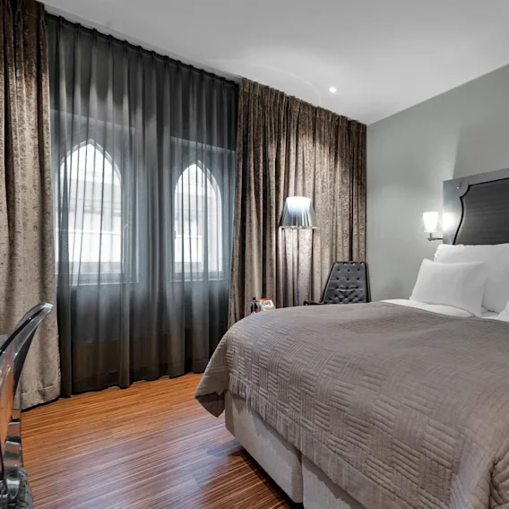 A neatly made double bed with gray and white bedding, placed beside a small nightstand and chair, is illuminated by soft wall lights in a modern, well-furnished bedroom.