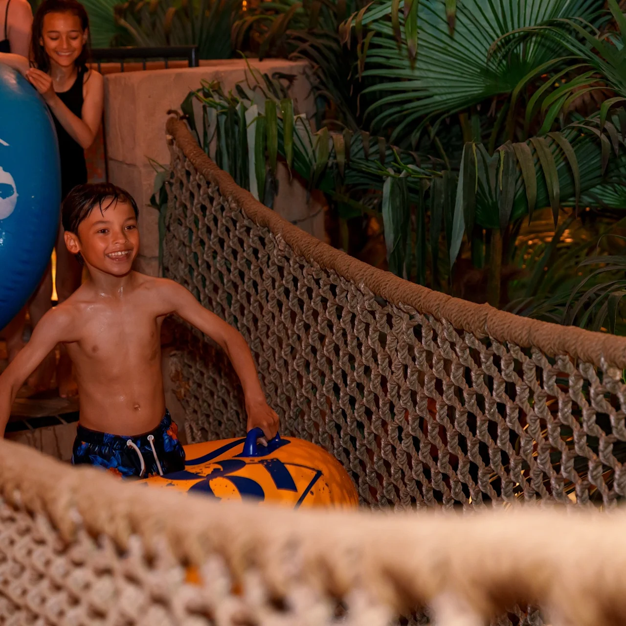 A boy sits excitedly on an inflatable tube, smiling. He is on a rope bridge surrounded by lush greenery. Another person behind holds a large blue inflatable ring.