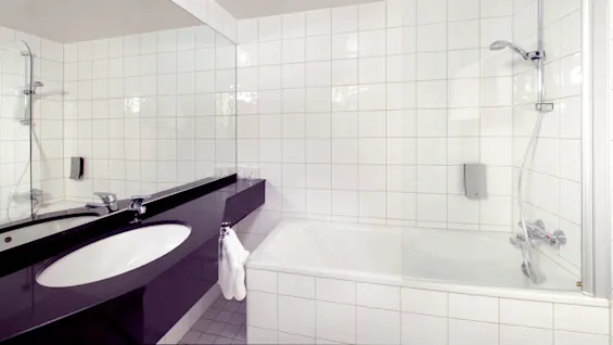 A clean bathroom with a white tiled wall, a large mirror above a black countertop with a sink, and a bathtub with a shower.