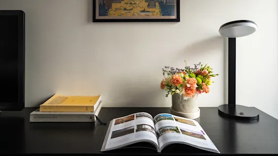 An open book lies on a dark desk with a vase of colorful flowers and a modern lamp. "THE DESIGN HOTELS BOOK" is stacked next to two yellow books. A framed cityscape above provides a backdrop.