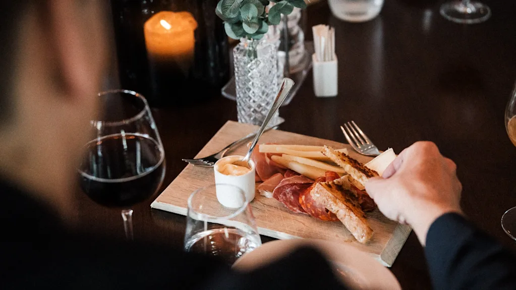 En person nyter et forrettsfat med spekemat, ost og brød, akkompagnert av vin i en koselig, levende lyssetting.