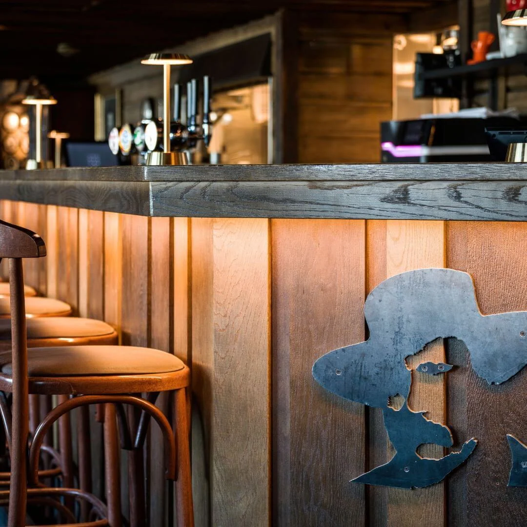 Bardisk med træstole arrangeret under varm belysning; baren har en metalsilhuet af en cowboy. Omgivelserne er hyggelige og dæmpet oplyst, med hylder med flasker i baggrunden.