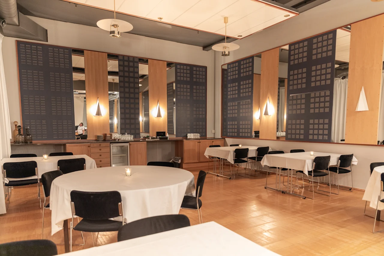 Tapahtumatila, jossa pyöreät ja suorakaiteen muotoiset pöydät on katettu valkoisilla liinoilla ja valaistu kynttilöillä, odottaen vieraita. Henkilö on taustalla.