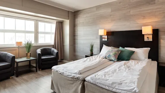 A neatly made double bed with white linens and decorative pillows is placed against a wood-paneled wall, flanked by lamps. A seating area with chairs and a table is by the window.