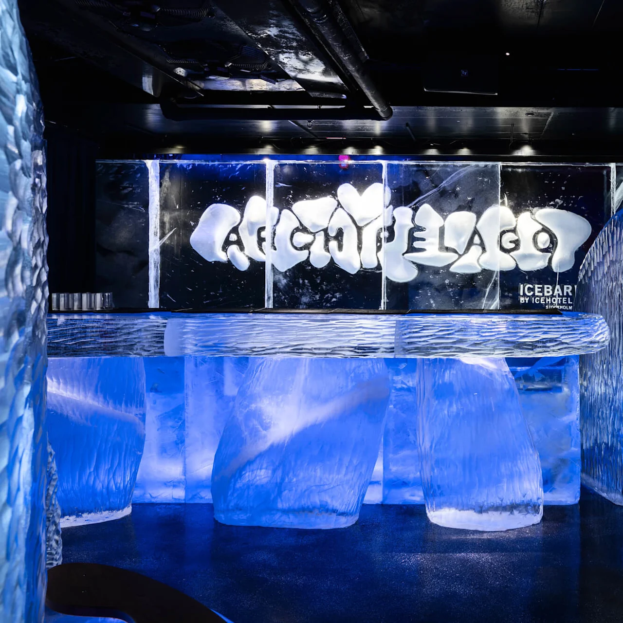 Ice bar, crafted entirely from ice, glows blue under lights. The counter and walls are transparent, with icy textures. Text: "ARCHIPELAGO" and "ICEBAR BY ICEHOTEL STOCKHOLM."