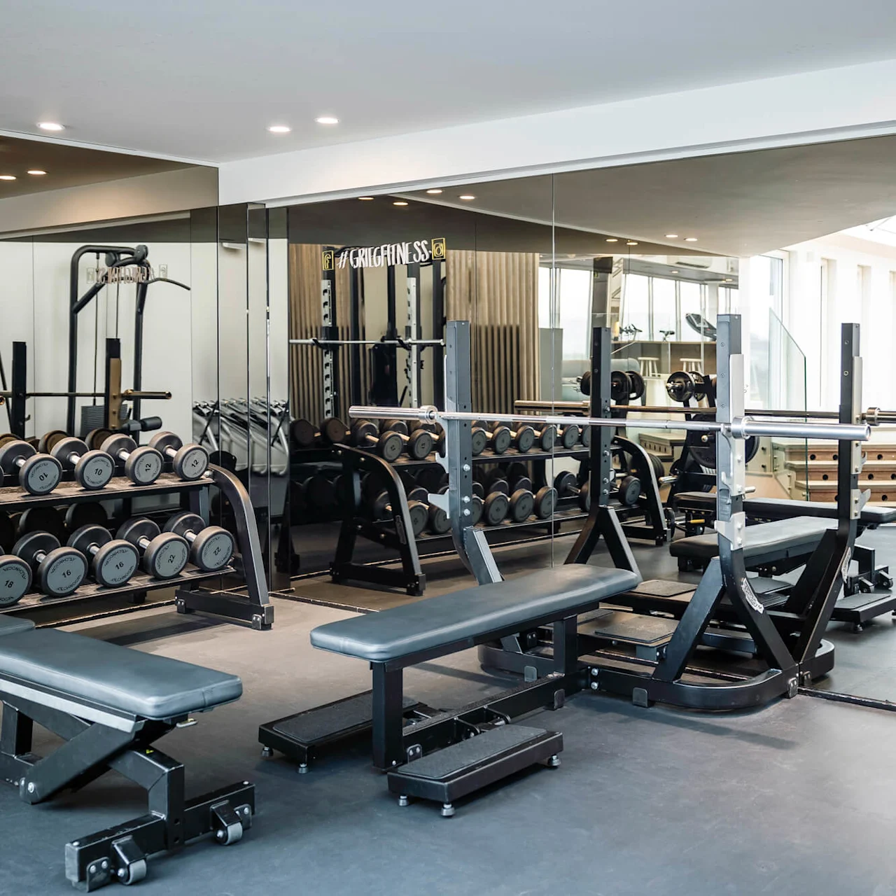 Treningsstudio med benker og en knebøyrack omgitt av manualer og speil; trapp synlig. Teksten "#GriegFitness" på speilet.