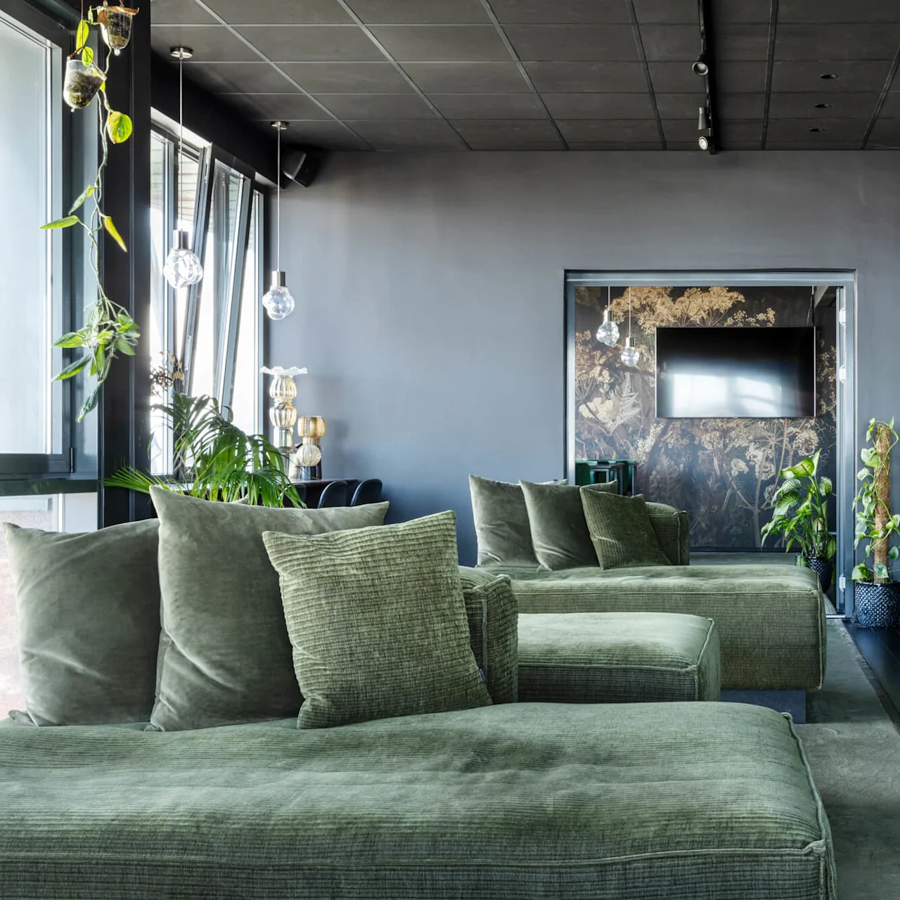 Large green sofas sit in a modern room with glass pendant lights and plants, under a dark ceiling. A wall-mounted TV faces a bar area with stools and more seating.