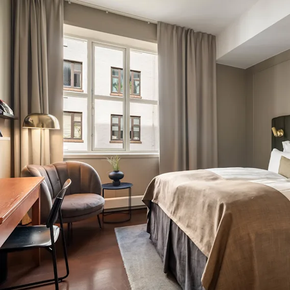A stylish bedroom with neutral tones, a large bed, desk, chair, and a window showing the exterior of a building.