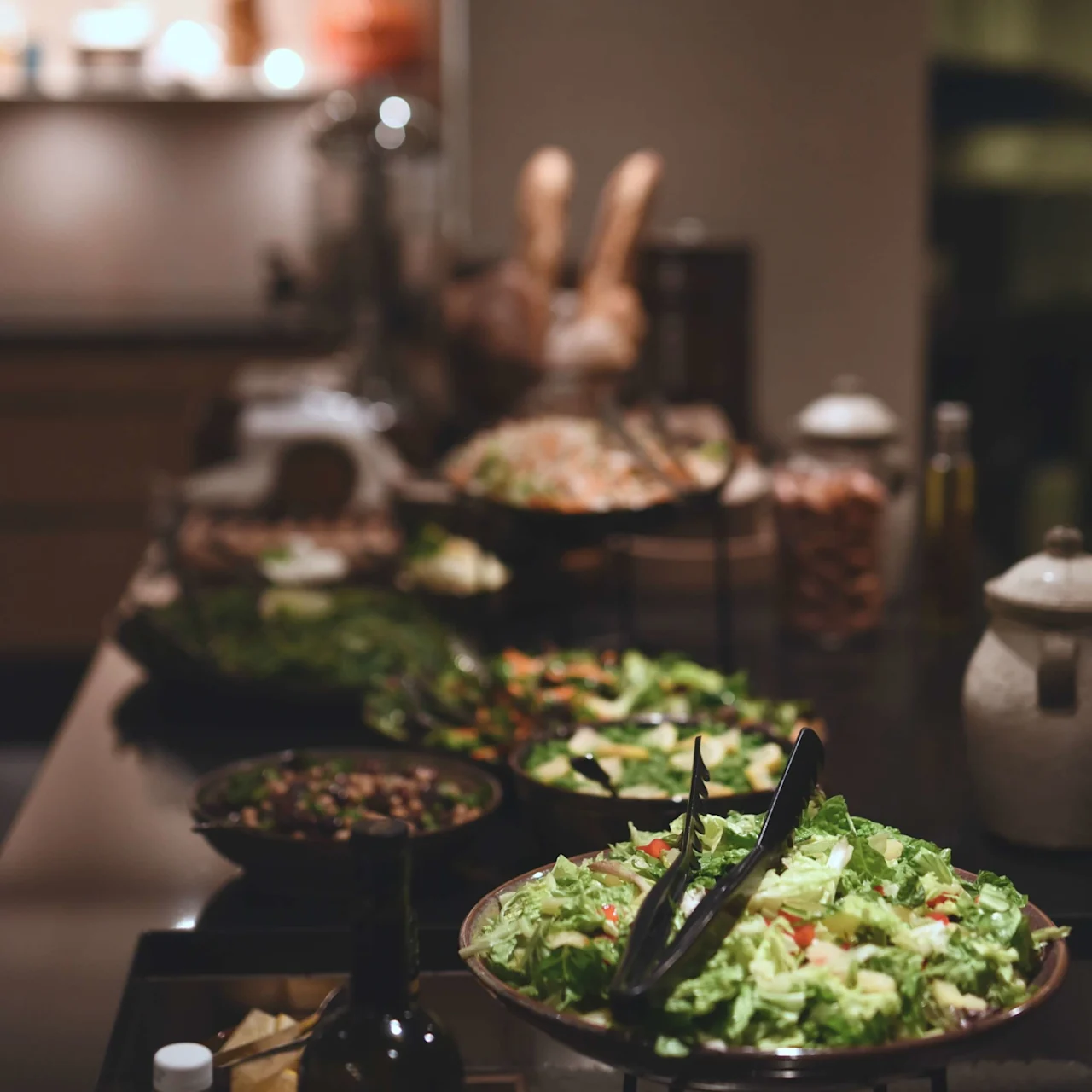 Skålar med färska sallader arrangerade på ett bord, med bladgrönsaker och färgglada grönsaker. I bakgrunden skapar olika sorters bröd och ett mjukt upplyst kök en varm atmosfär.