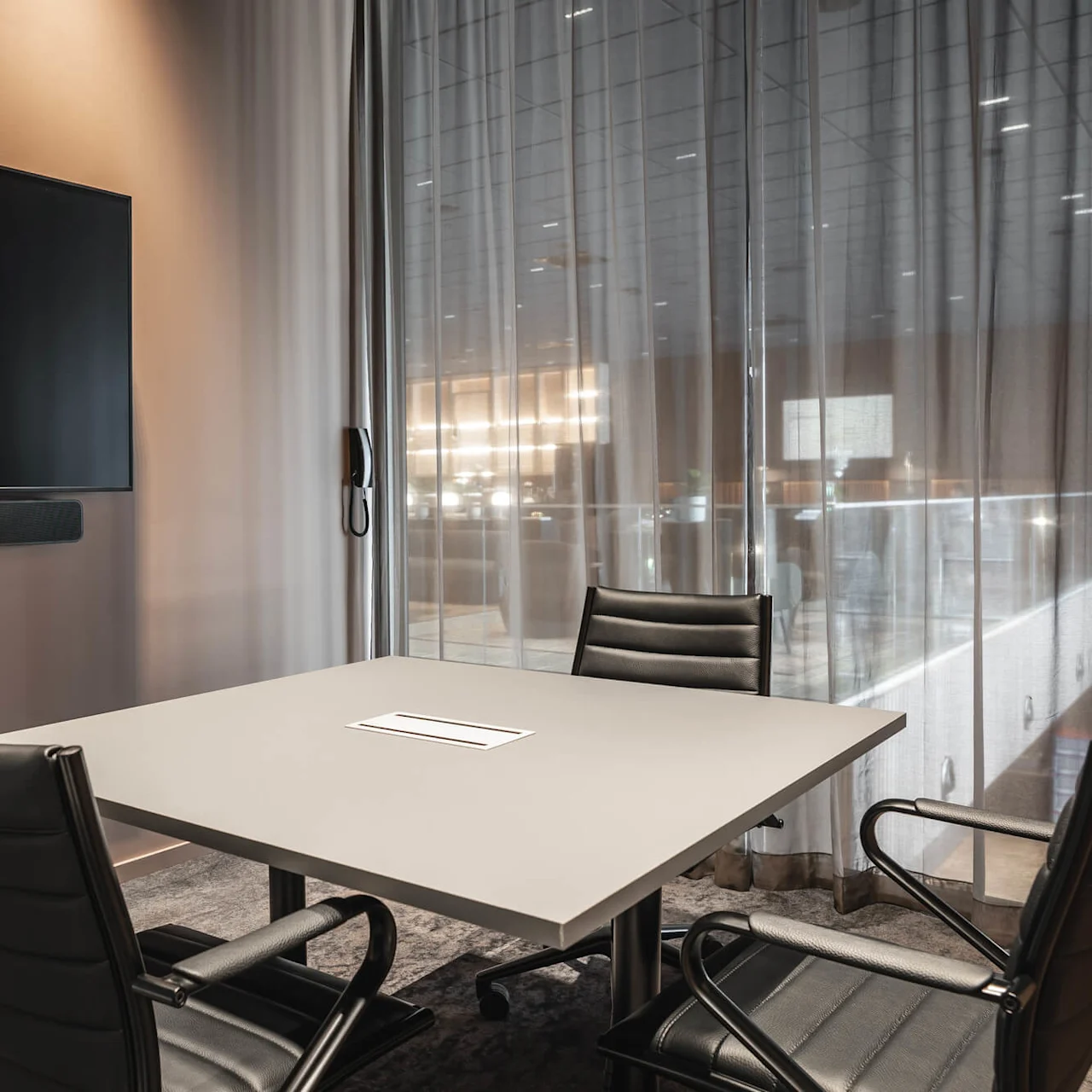 A conference room features a rectangular table surrounded by four black chairs. A large screen is mounted on the wall, with sheer curtains revealing a softly lit area beyond.