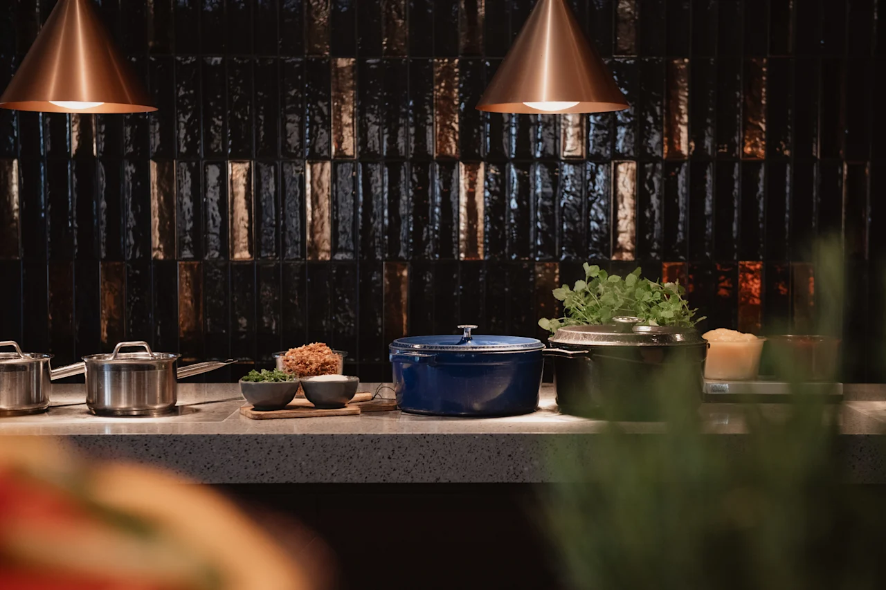 En moderne kjøkkenbenk med kokekar, ferske ingredienser i skåler og planter, belyst av stilige pendellamper.